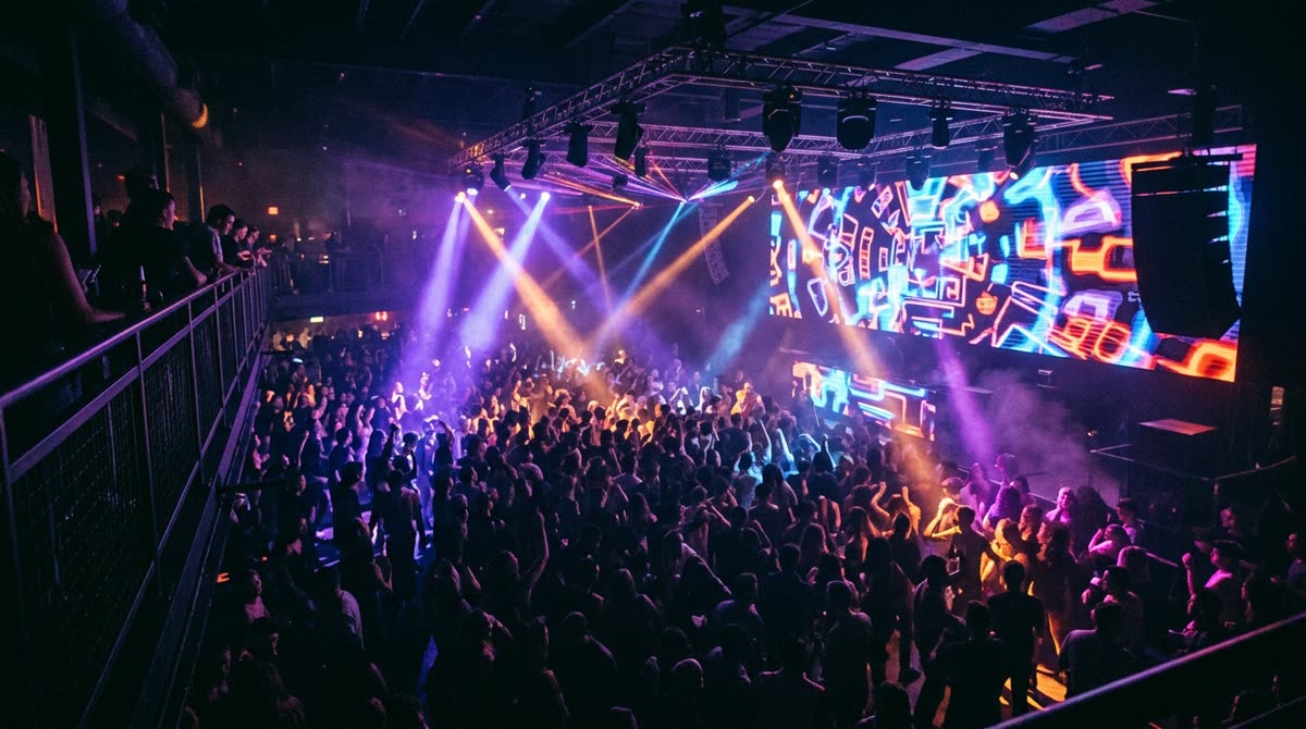 A crowded nightclub dance floor seen from a balcony with light beams cutting through haze.